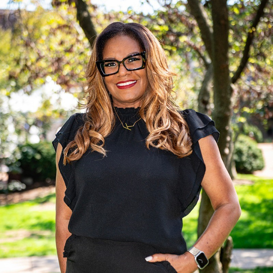 Image of Knieba Jones-Johnson, a Black woman standing outside with long, blond hair and a bright smile with red lipstick. She is wearing large rimmed, black glasses and a black blouse with short sleeves.