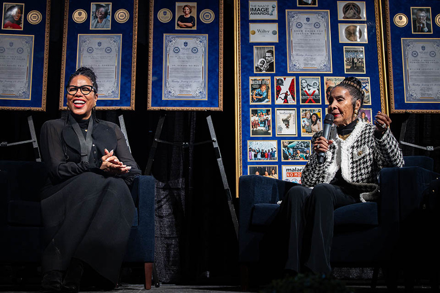 two Black women sitting on chairs on stage in front of a wall of awards 