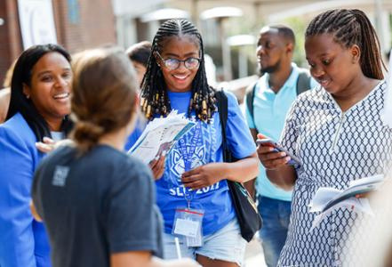 Image of multiple students an an event, with a clear view of a young Black woman with her hair in locs, wearing glasses and a blue SLU t-shirt, and sharing a large smile. 