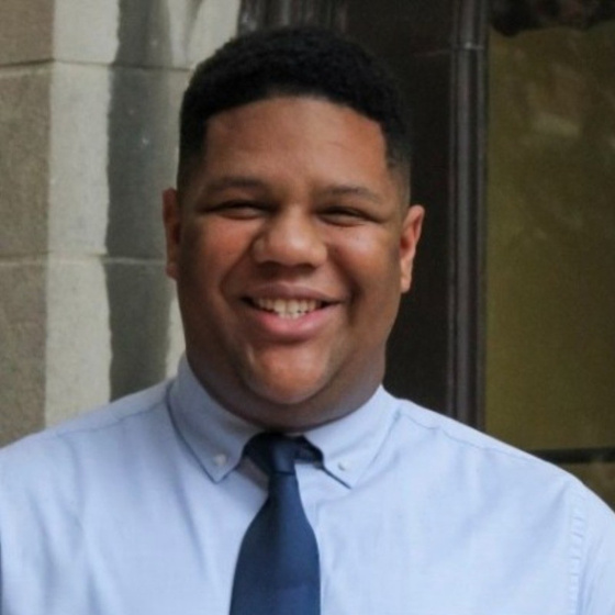 Image of Ryan Tisdale, a young Black man and scholar, who has short styled hair with clean lines and a wide smile. Ryan is wearing a light blue collared shirt with a navy blue tie. 